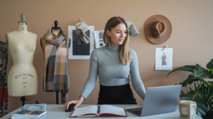 Eine Frau im grauen Rollkragenpullover arbeitet an einem Schreibtisch mit Laptop und aufgeschlagenem Notizbuch. Hinter ihr hängen Schaufensterpuppen mit Schal, Inspirationsfotos und ein Hut an der Wand. Rechts steht eine Topfpflanze.