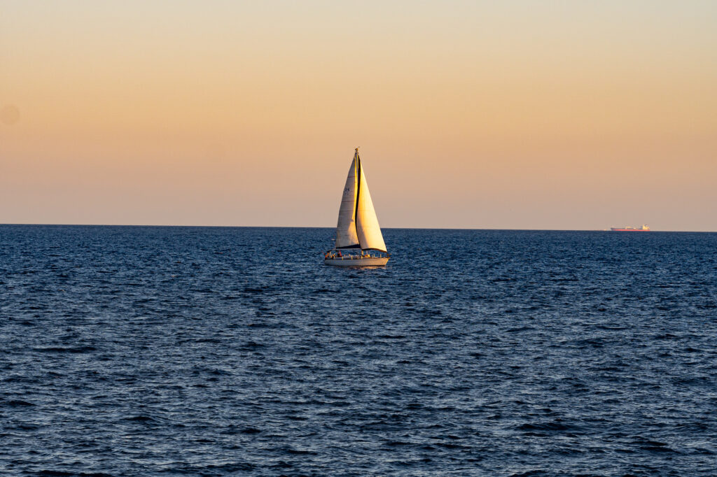 Ein Segelboot gleitet bei Sonnenuntergang unter einem klaren Himmel über ein ruhiges Meer. Der Horizont leuchtet in einem sanften Orangeton. In der Ferne ist ein Frachtschiff zu sehen.